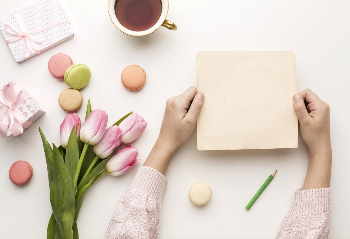 Woman's hands writing in a greeting card next to a cup of tea and a small vase of pink flowers
