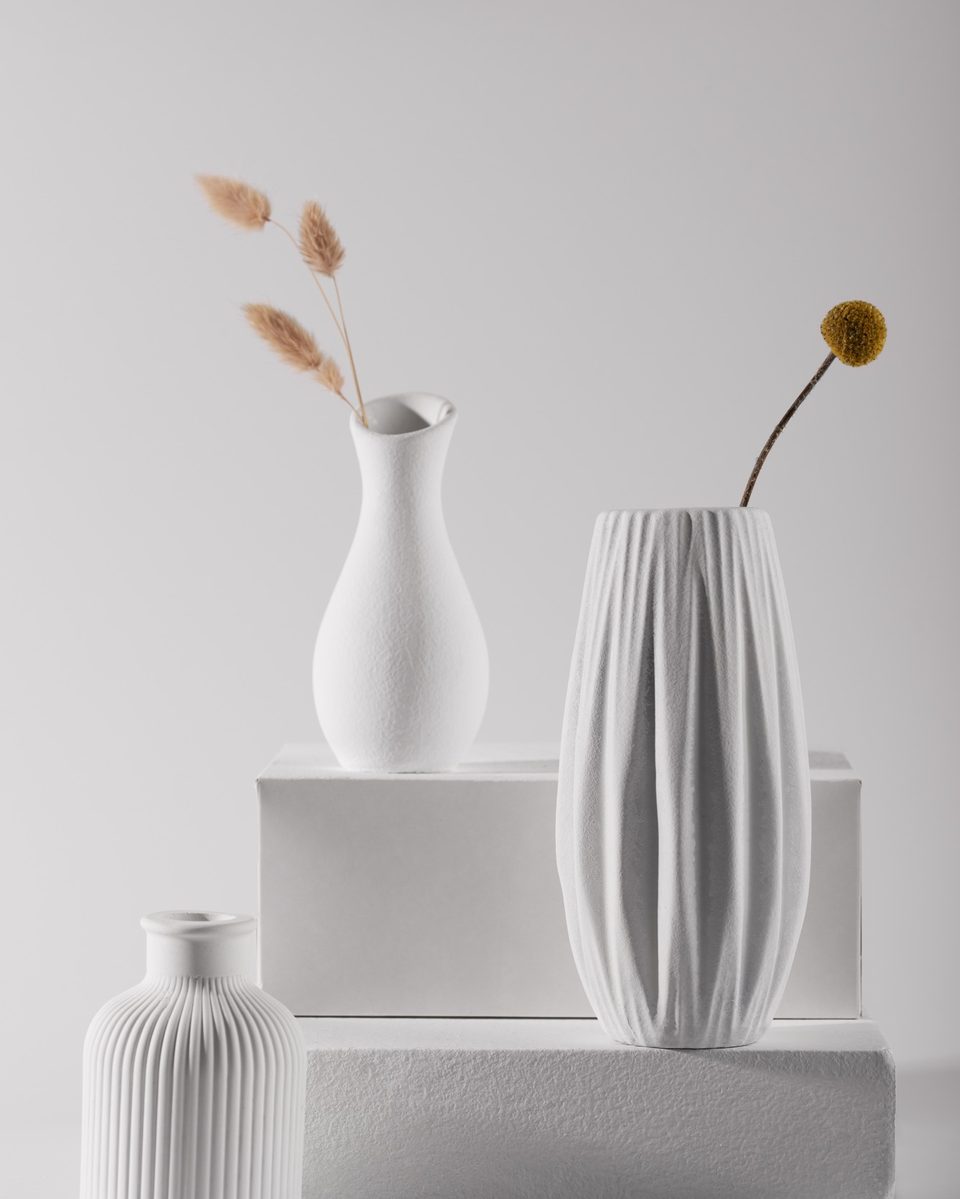 Group of white ceramic vases in different shapes with dried stems on a white plinth