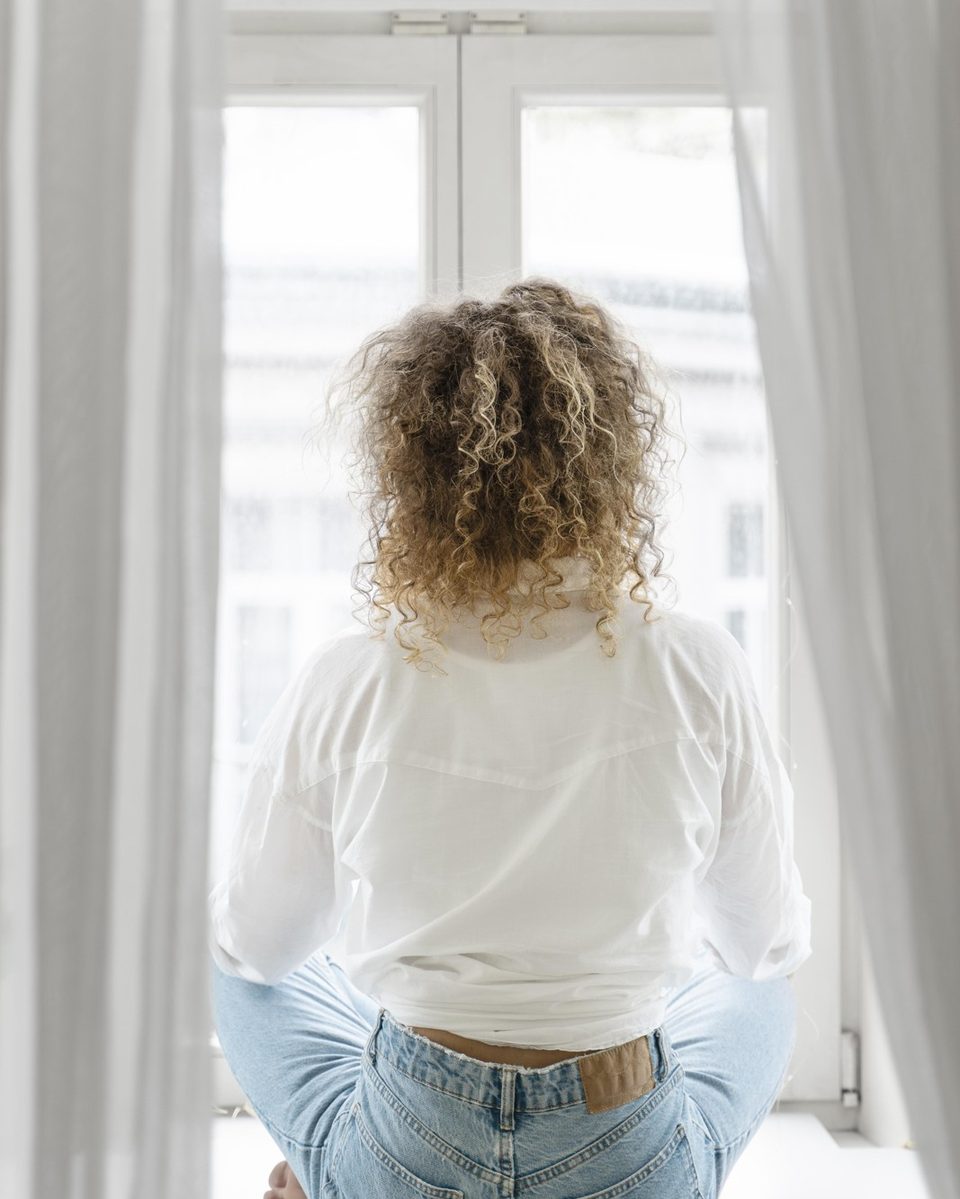 Woman seen from behind sitting cross-legged on a beige cushion by a bright window in soft morning light