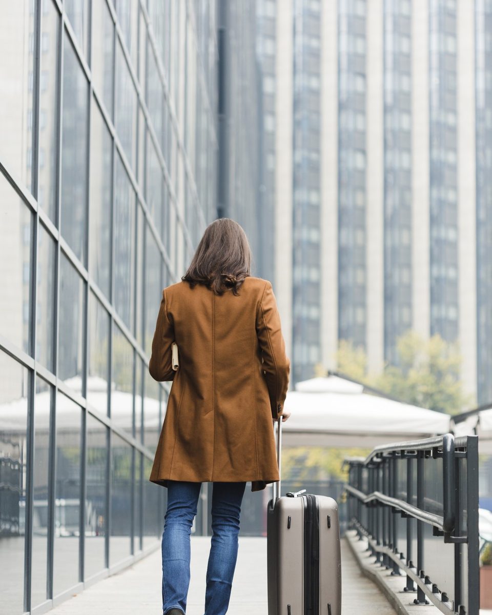 Woman seen from behind walking through a bright airport with a cream carry-on suitcase and a beige trench coat
