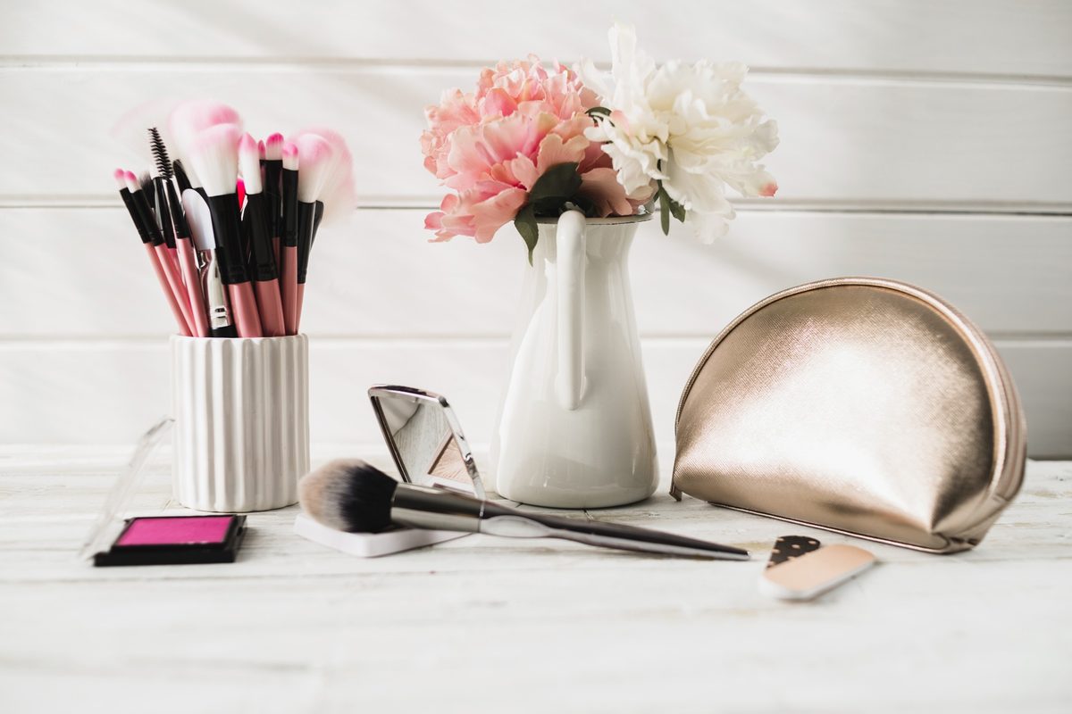 Pink makeup brushes in a cup, a rose gold makeup bag and flowers in a white vase