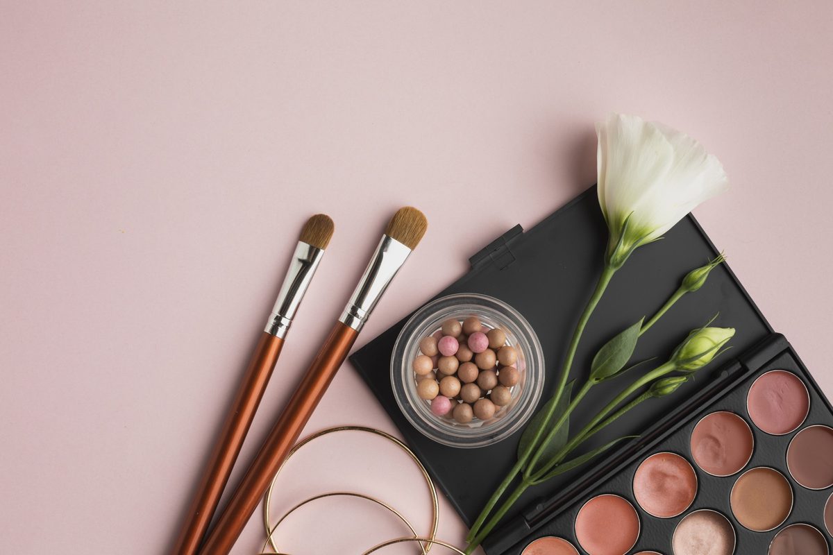 Makeup palette, two brushes, bronzing pearls and a white flower on a pink background