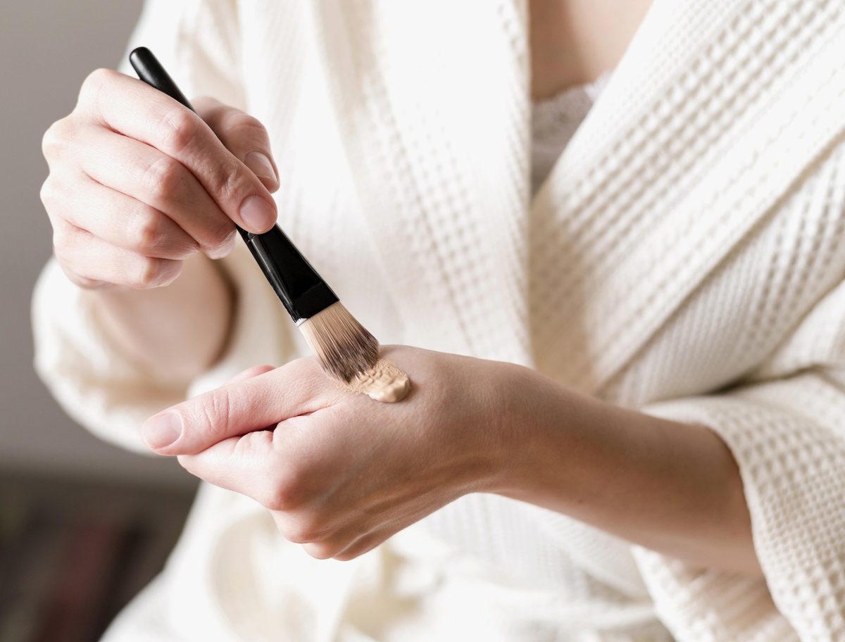 Woman in a cream robe testing a foundation shade on the back of her hand with a brush