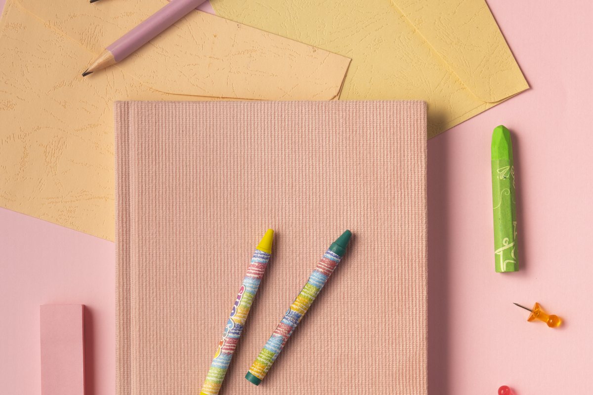 Blush pink notebook with pastel crayons and soft yellow paper on a pink background