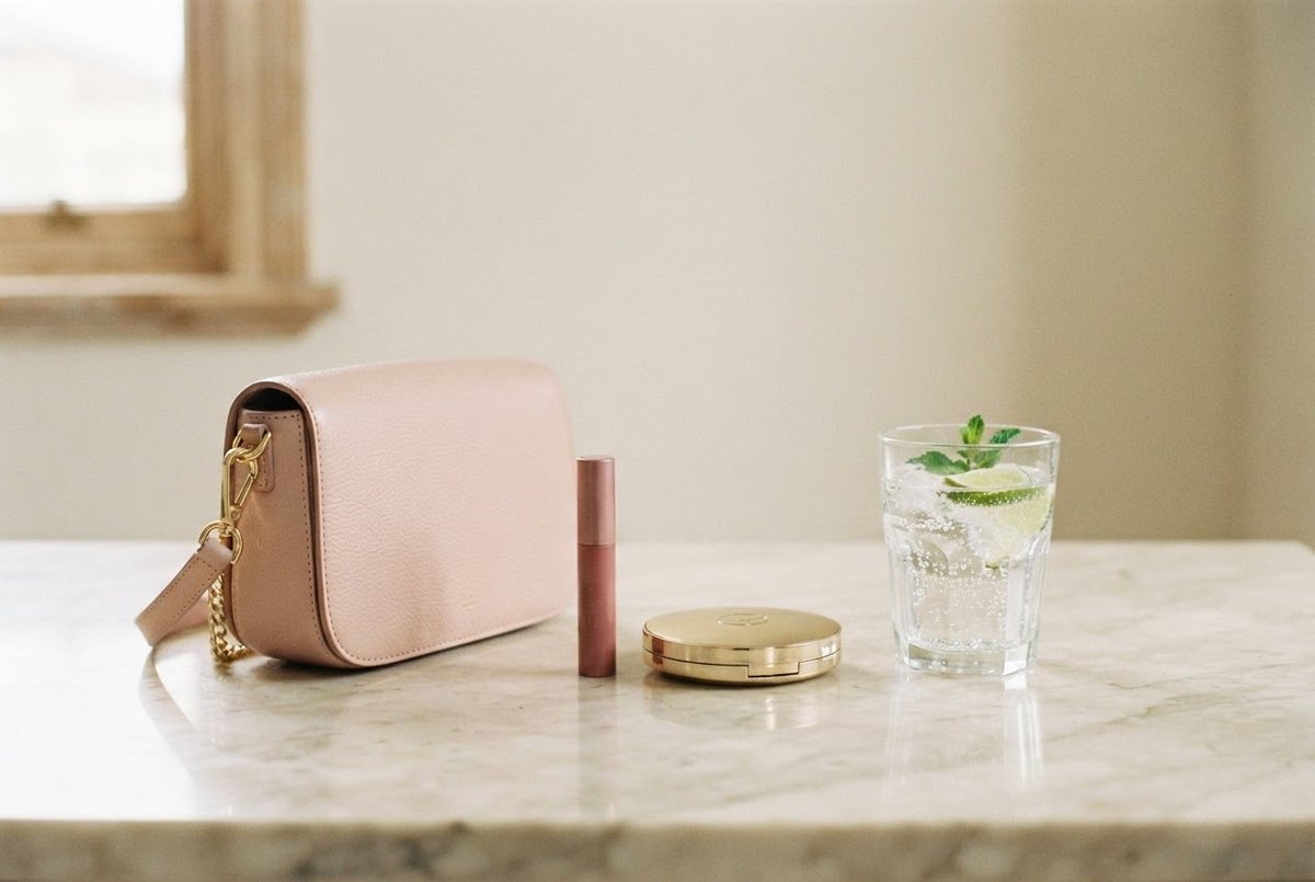 Blush crossbody bag, a lip tint, a gold compact and a glass of sparkling water on a cream marble café table