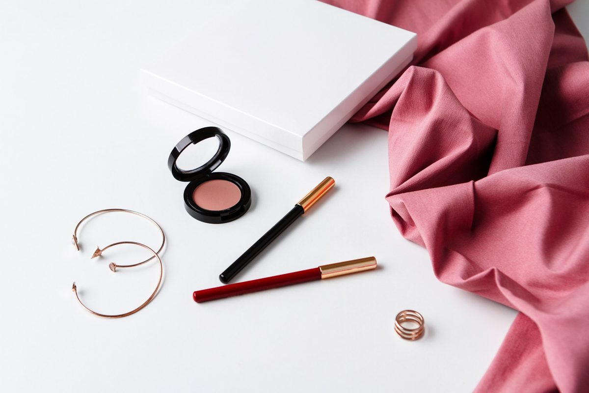 Flat lay of lipstick, blush compact, eyeliner and gold earrings with a pink silk fabric
