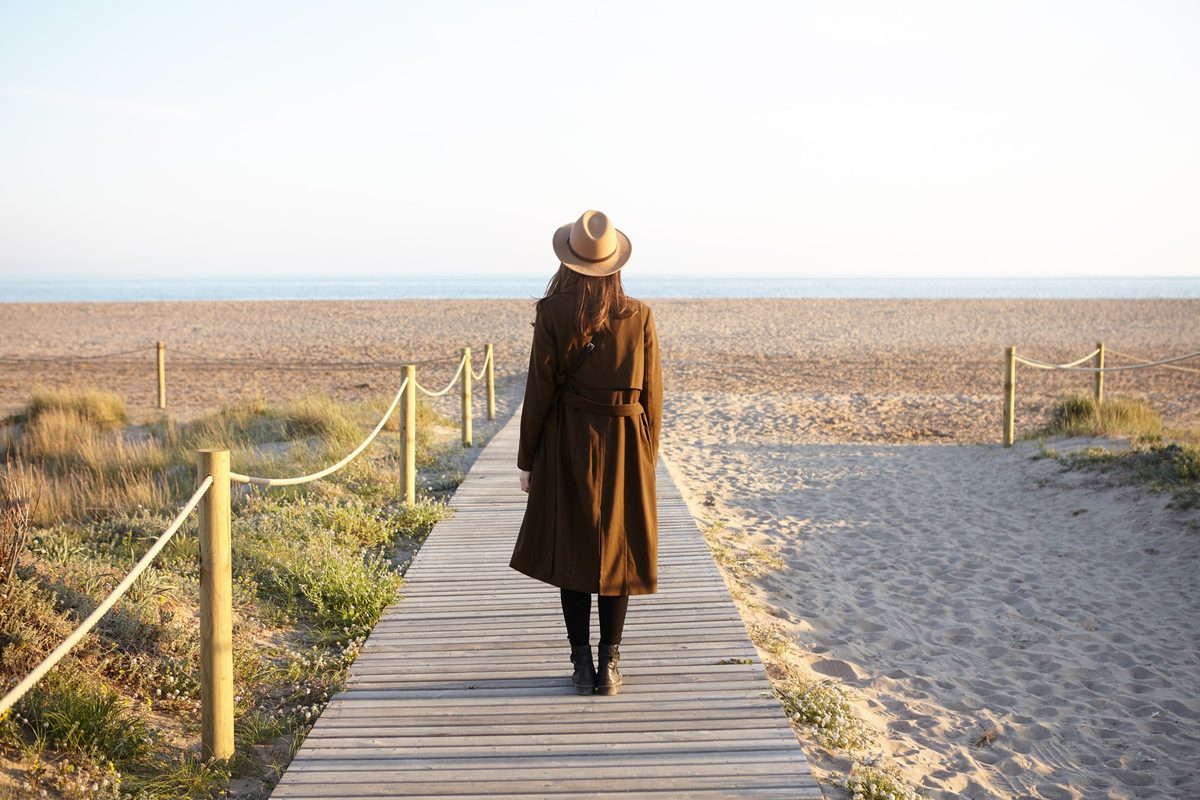 Woman in a hat and long coat seen from behind walking along a wooden boardwalk by the sea