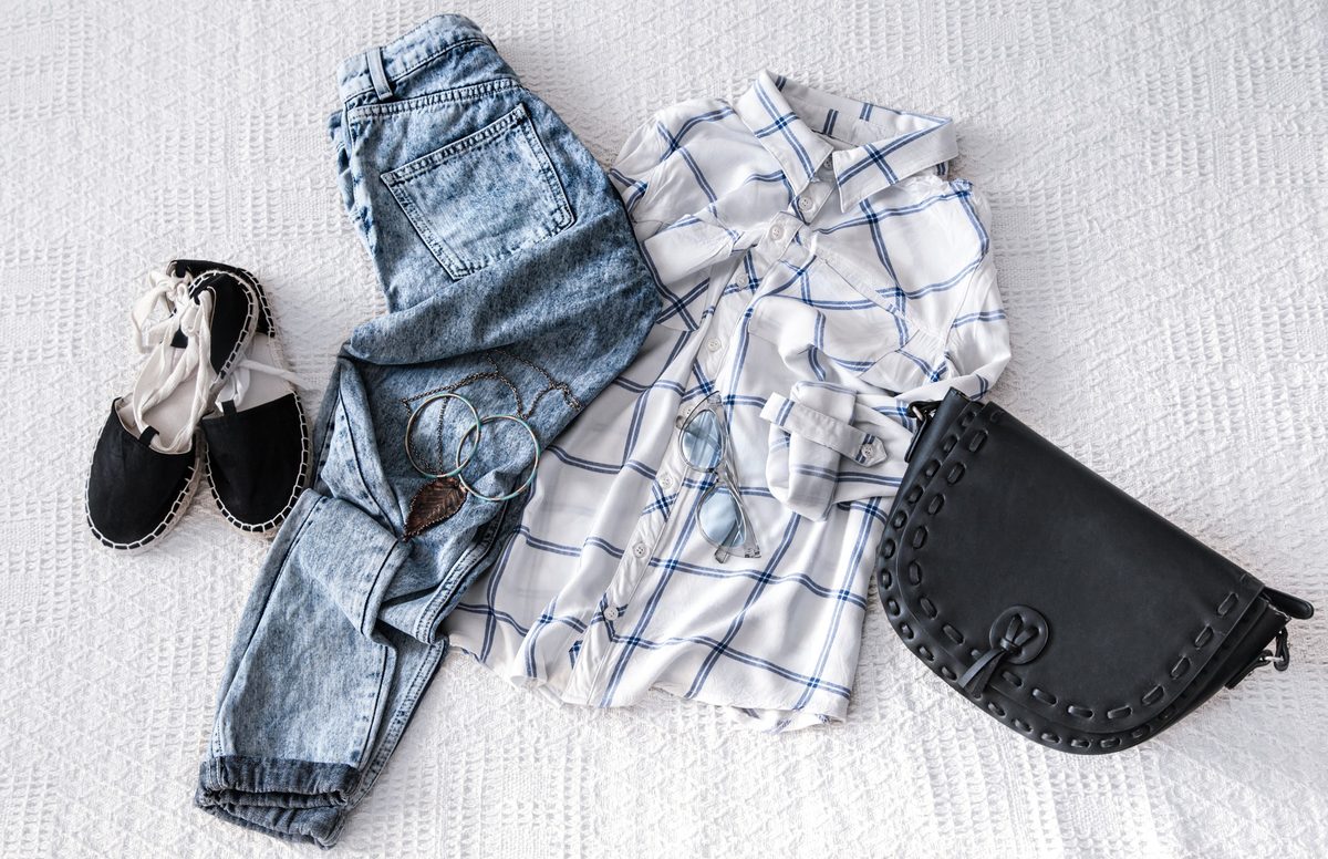 Women's checked shirt, light jeans, espadrilles and a dark clutch laid flat on white