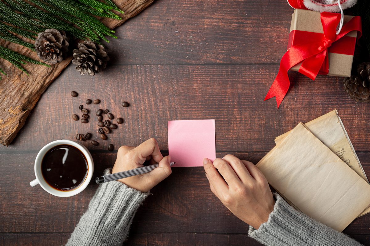 Hands writing a little love note on a pink paper beside a cup of coffee and a wrapped gift