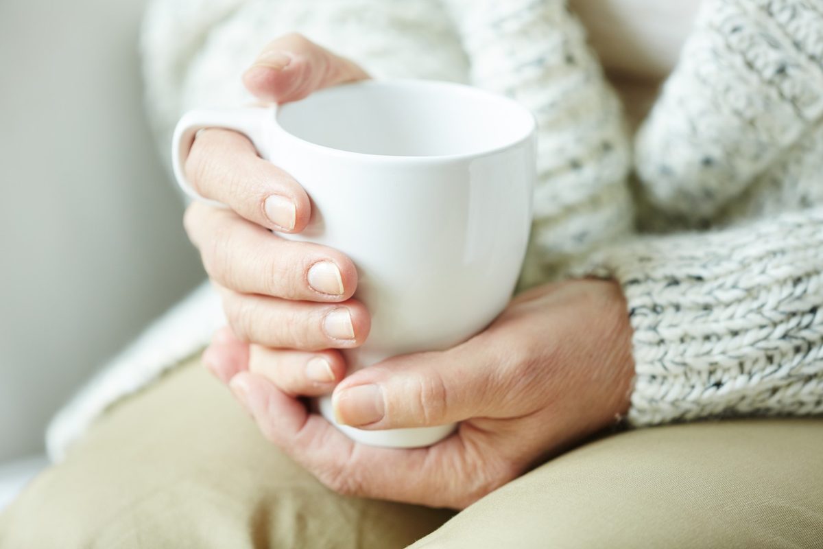 Cropped view of a woman with soft gray hair in a lavender knit sweater holding a white mug, face not visible