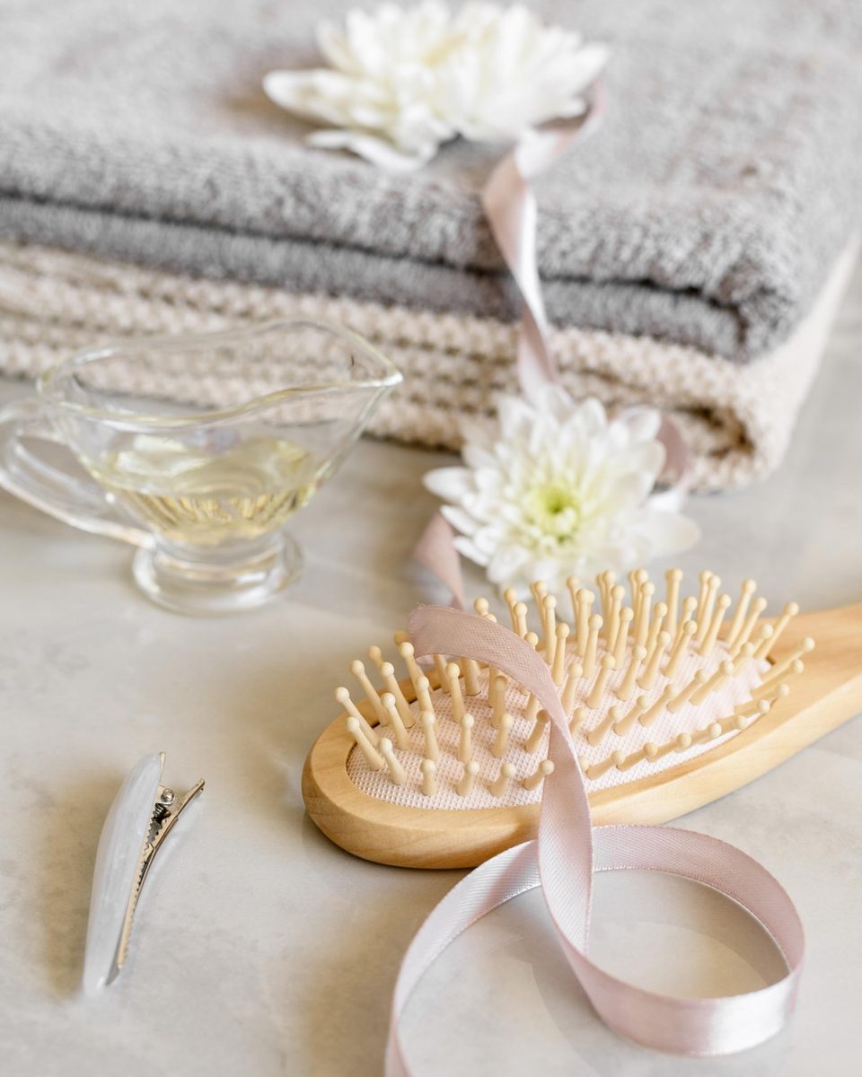 Flat lay of a wide-tooth comb, cream silk scrunchies, a lavender shampoo bottle and a folded white towel on a marble surface