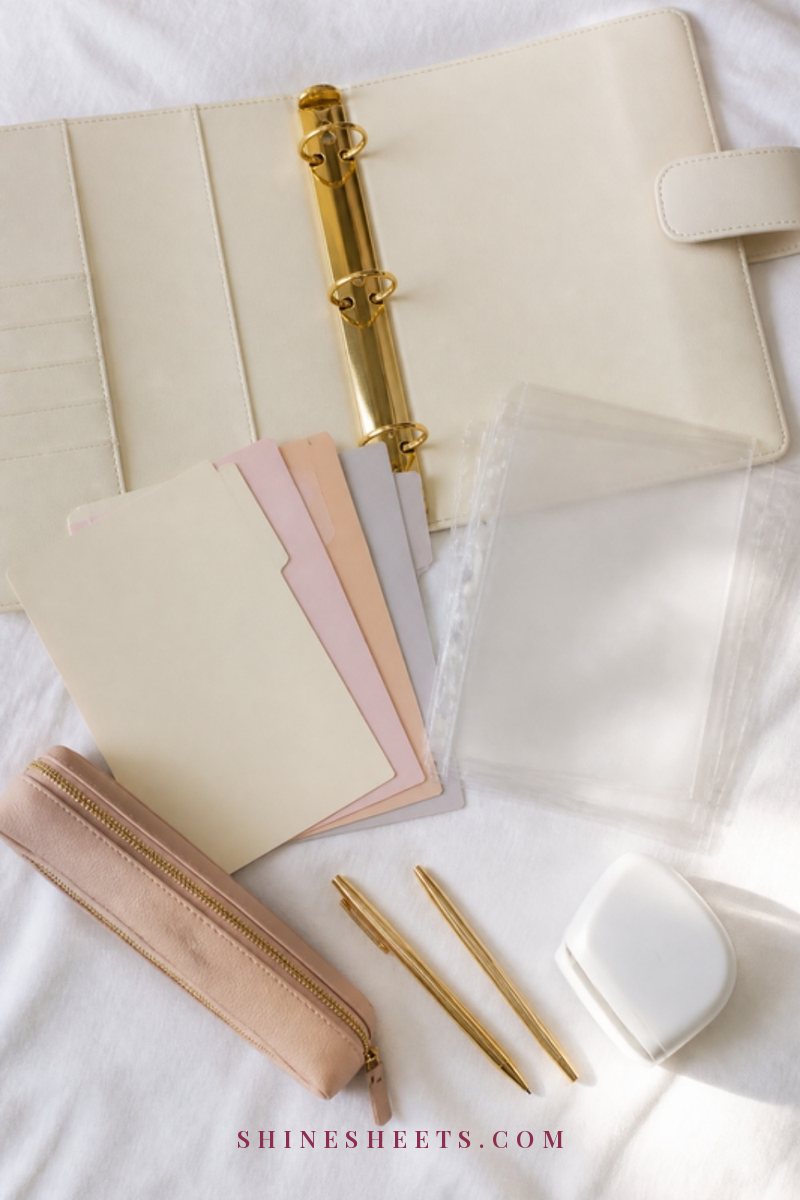 Flat lay of a cream binder, pastel tab dividers, clear sheet protectors, a blush zipper pouch and gold pens on a white linen surface