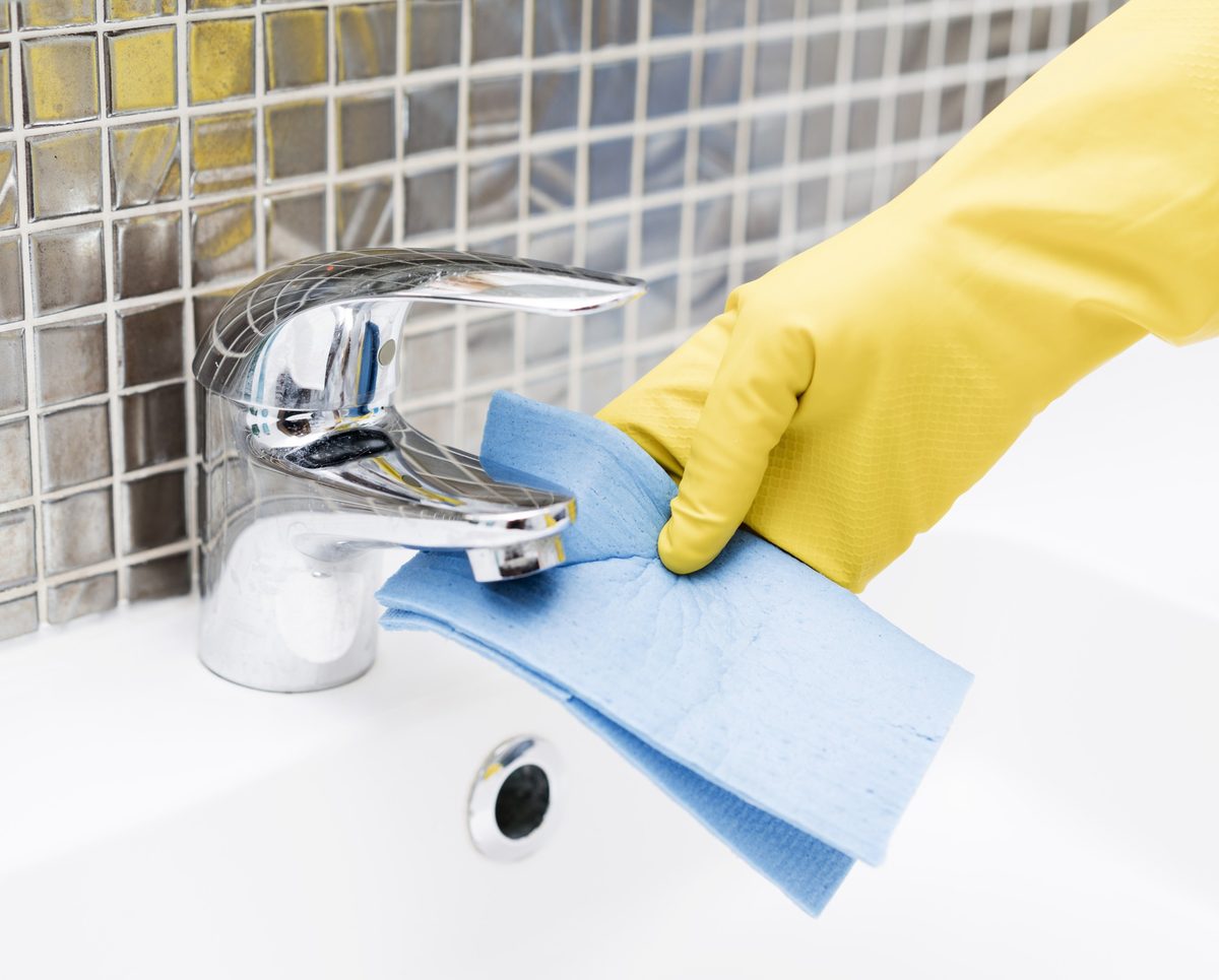 Hand in a yellow rubber glove wiping a bathroom faucet with a blue cloth