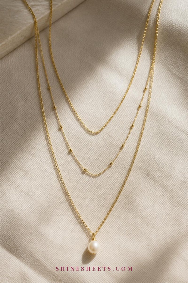 Three fine gold necklaces laid in a cascading V on cream linen, spaced evenly with a small pearl pendant at the bottom