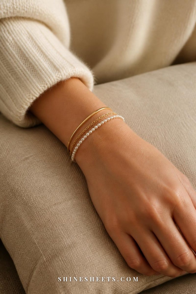Woman's wrist on a beige linen cushion wearing a slim gold bangle, a fine chain bracelet and a thin pearl bracelet