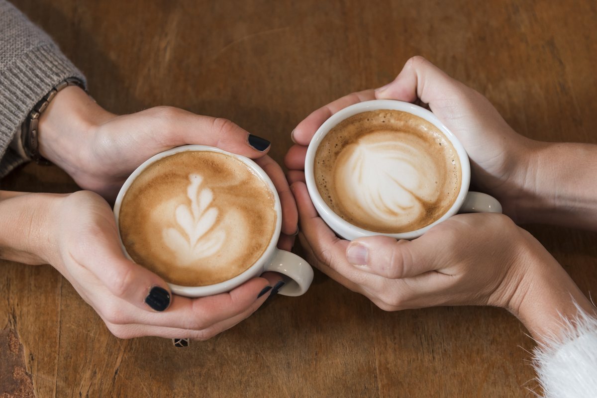 Two friends holding latte cups with heart foam art on a wooden table