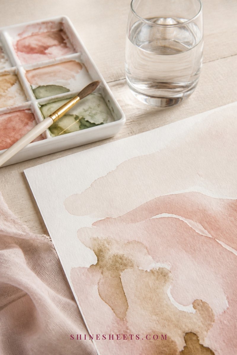 A watercolor palette, a soft brush and a sheet of paper with loose blush and beige color washes on a cream wooden table