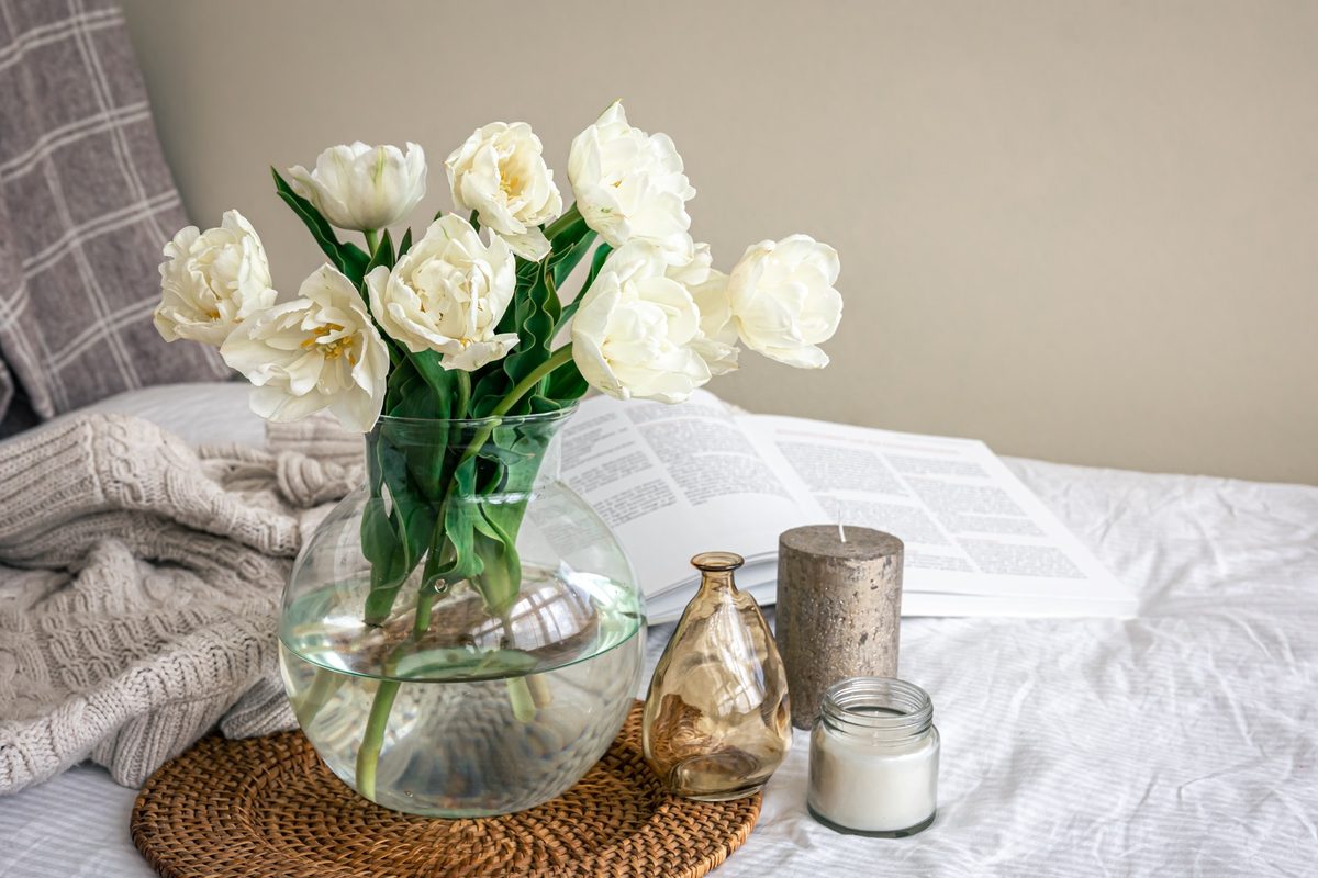 White tulips in a glass vase with candles and a knit throw on a white bed