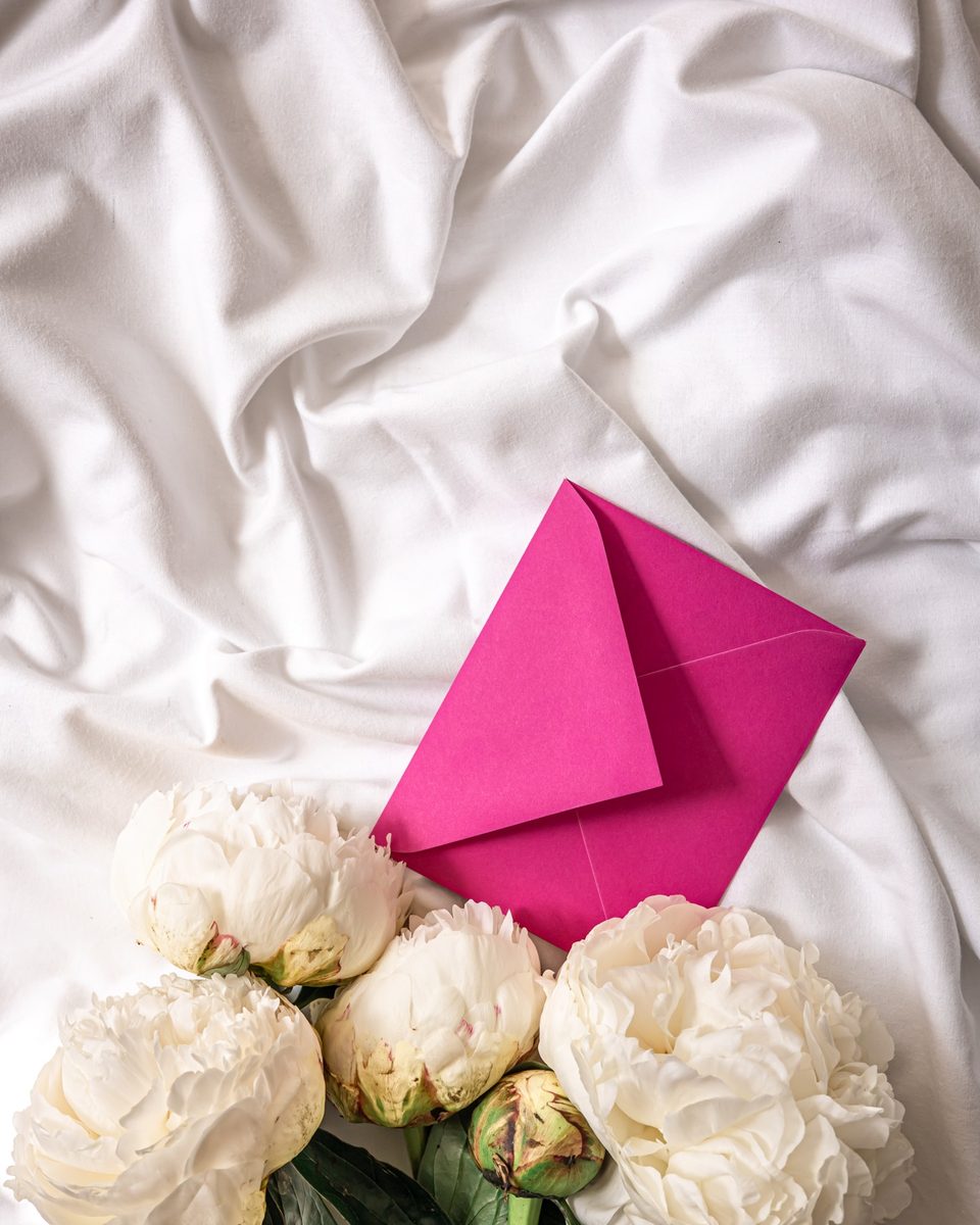 Pink envelope and white peonies on soft white sheets