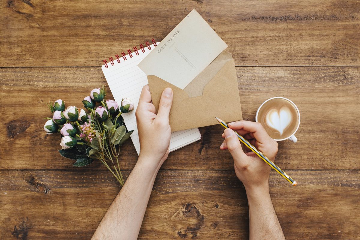Woman's hands writing a card with a pen next to a latte and berries on a wooden table