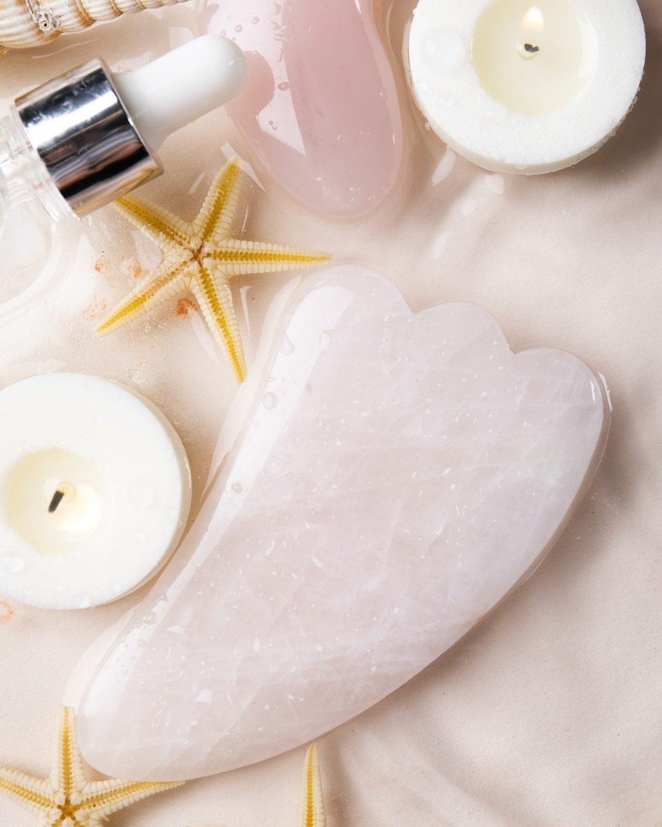Glass candle, a cup of herbal tea and a ceramic dish with a gua sha stone on a white nightstand in warm evening light