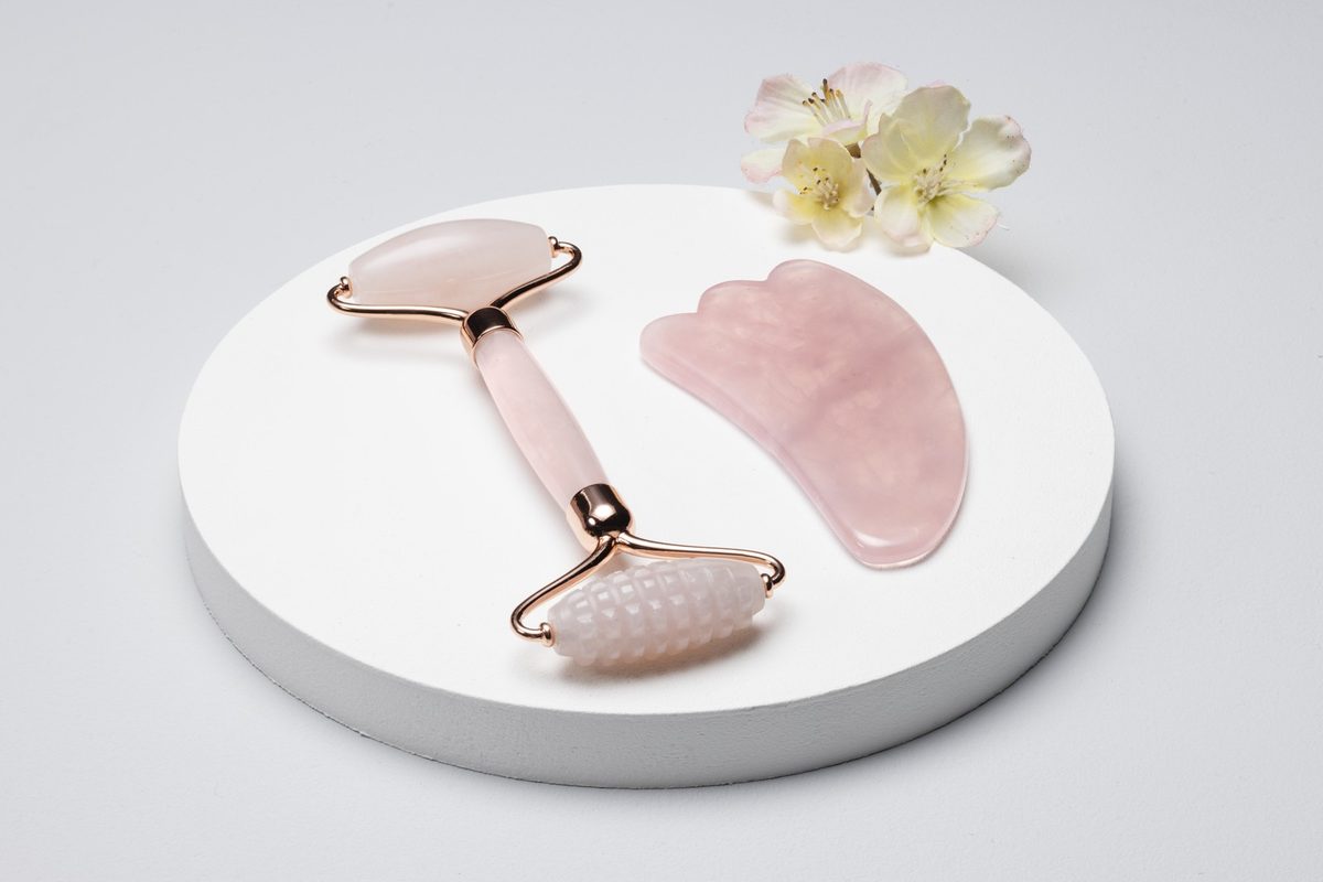 Rose quartz gua sha stone, a small facial oil bottle and a white towel on a cream marble bathroom counter