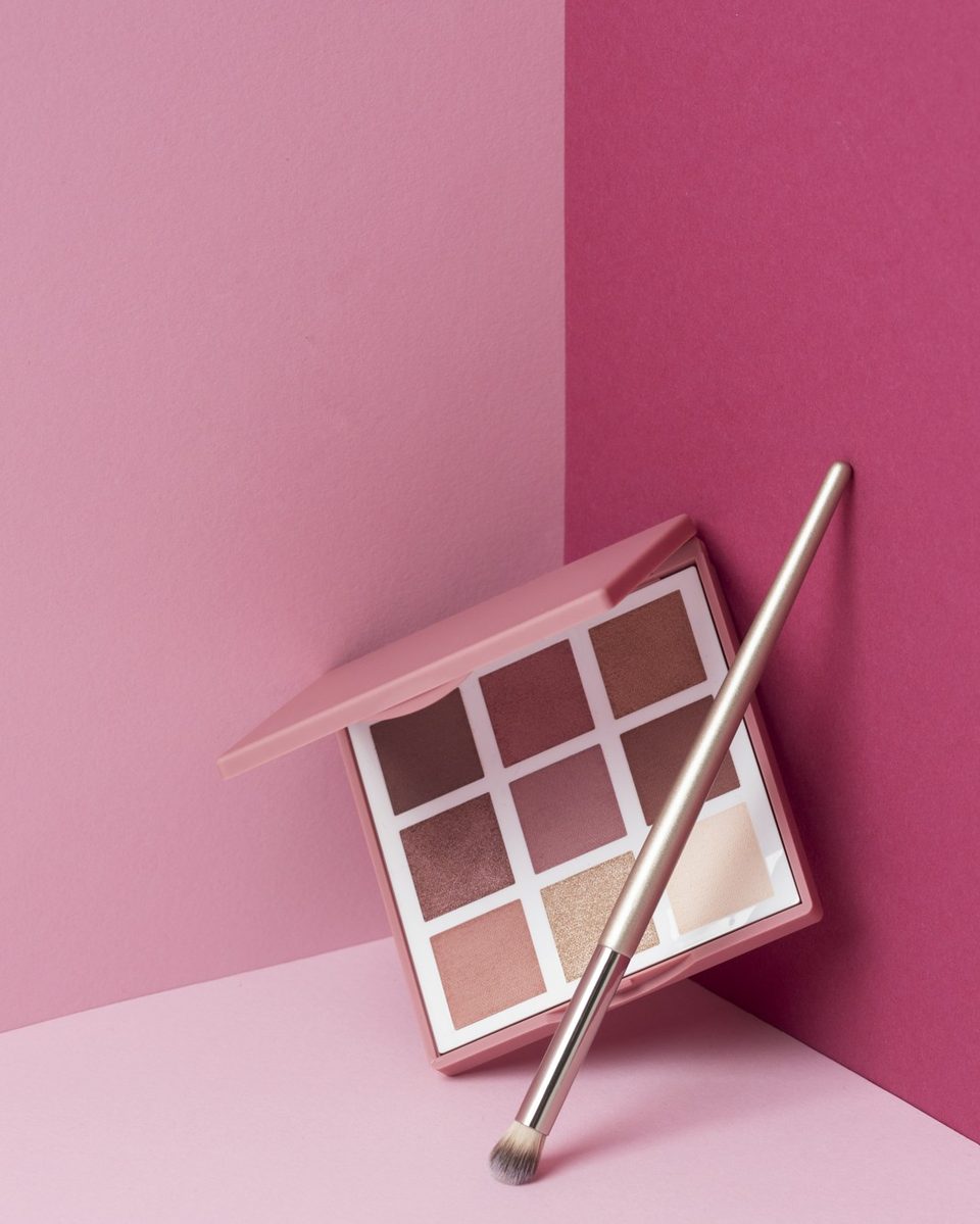 Rosy lipstick, a brow pencil and a small eyeshadow palette on a blush pink silk scarf beside a glass candle