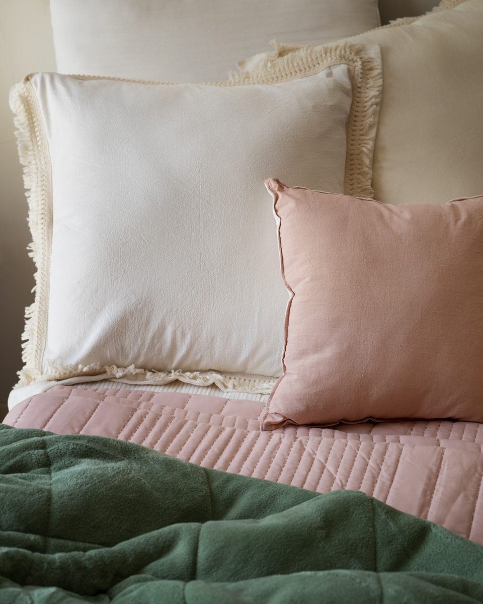 Neatly made bed with white linen, a blush quilt, gingham pillows and a knit throw at the foot of the bed