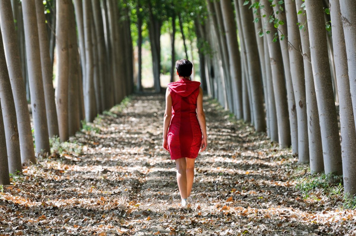 A woman seen from behind walking on a quiet tree-lined path in soft morning light, wearing a cream knit sweater