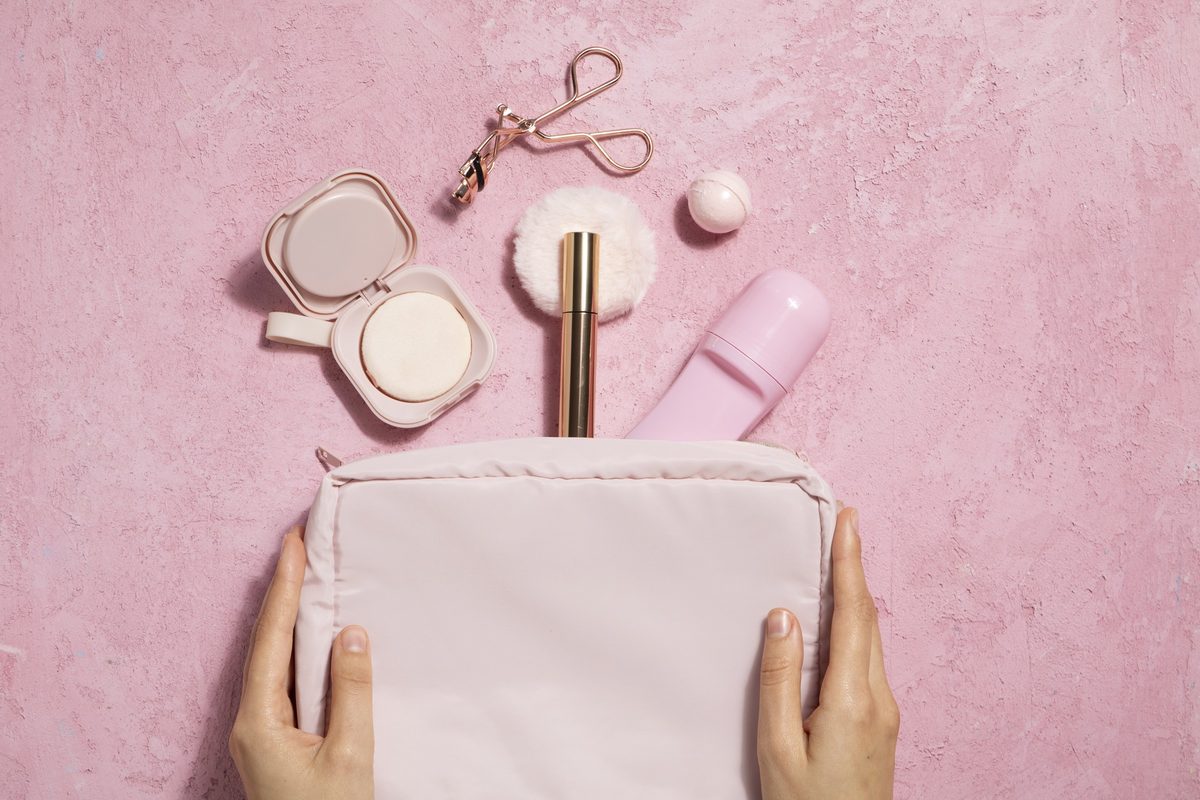Woman's hands holding a pink makeup bag below a compact, an eyelash curler and a lipstick