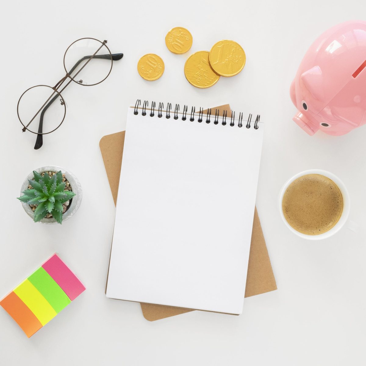 Blank notepad, glasses, coins, a coffee and a pink piggy bank on a white desk