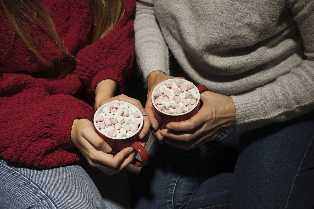 Couple in cozy sweaters holding mugs of hot cocoa with marshmallows