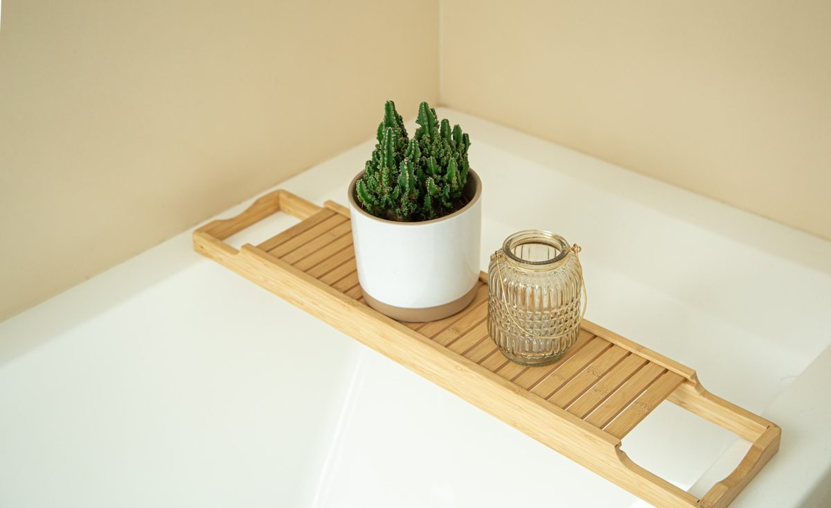 Wooden bath caddy with a glass candle, a white towel and a small bowl of bath salts across a white bathtub
