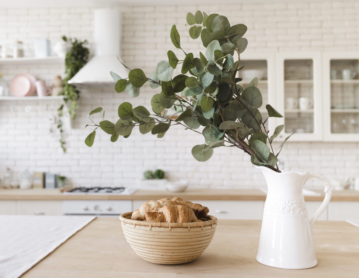 Clean kitchen counter with a cream kettle, a utensil crock, a wooden board and a small plant by a window with a linen curtain