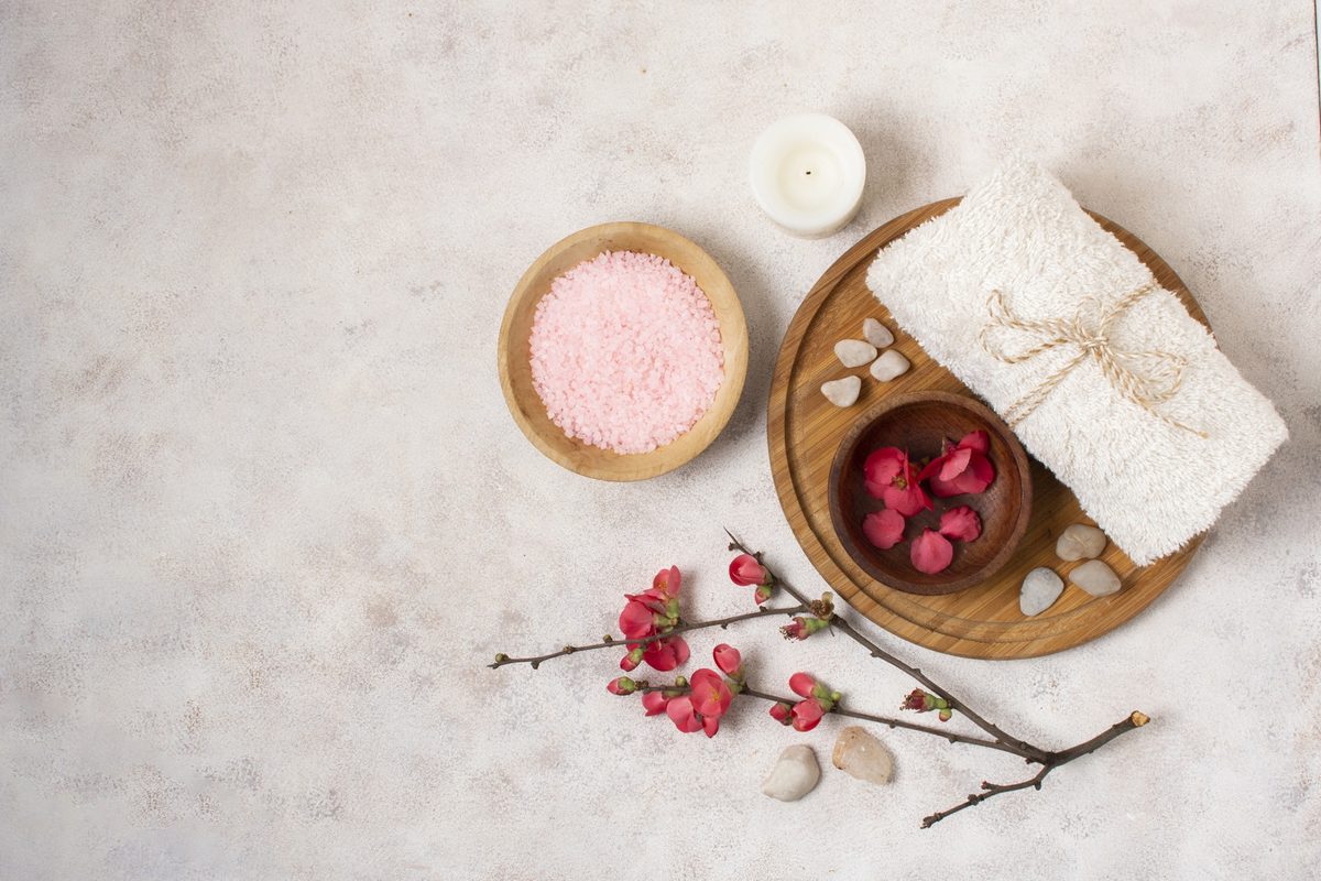 Pink bath salts, a rolled towel and rose petals arranged on a light surface