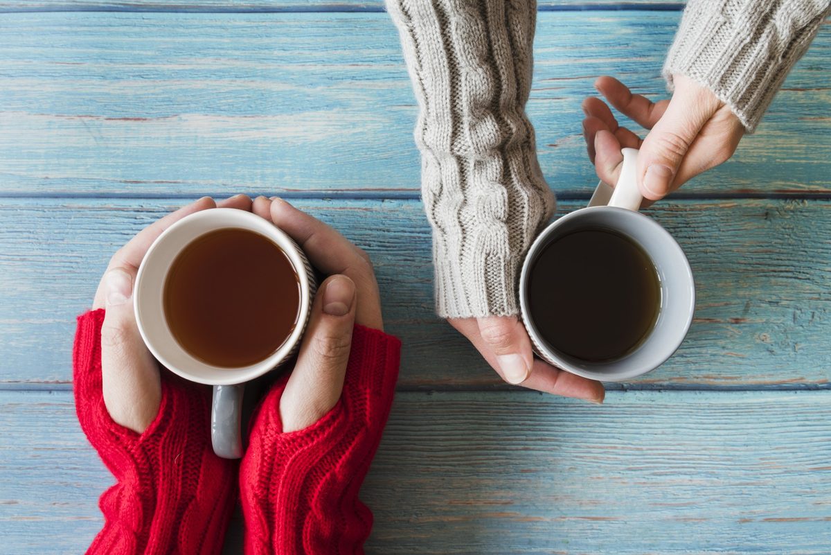 Two people in cozy sweaters holding mugs of tea on a blue wooden table