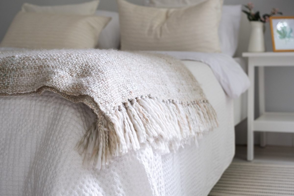 Neat bed with layered cream and blush linen bedding, a knit throw and soft pillows in a small sunlit bedroom