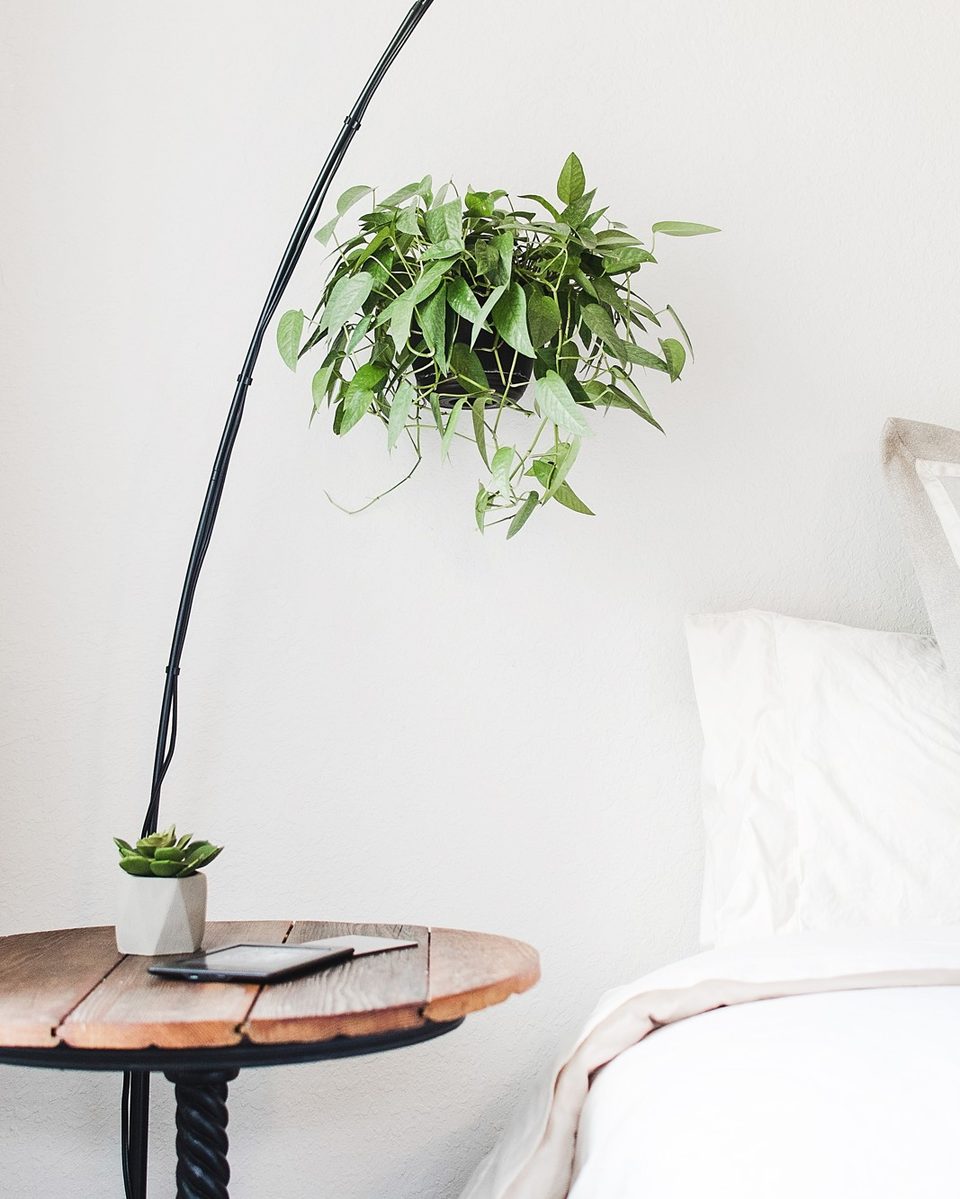 White floating shelf beside a bed with a small plant, a glass candle and a stack of books against a cream wall