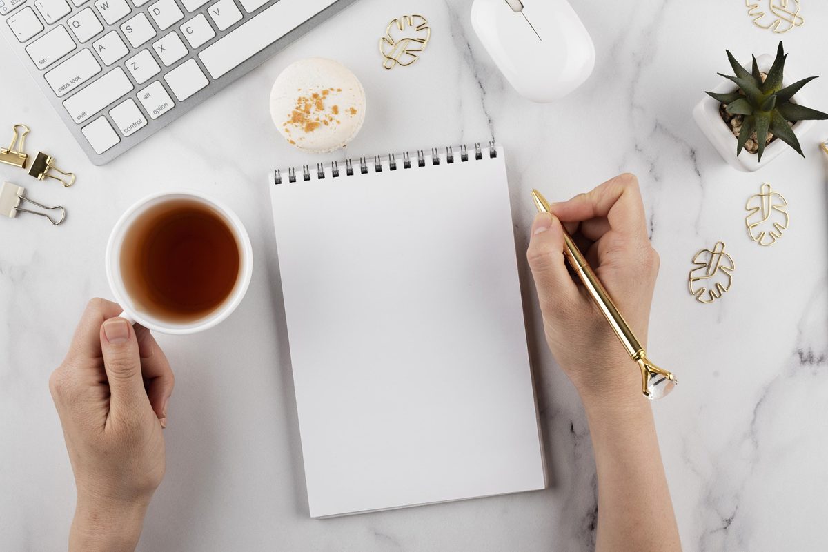 Woman's hand writing in a blank notepad next to a cup of tea on a marble desk