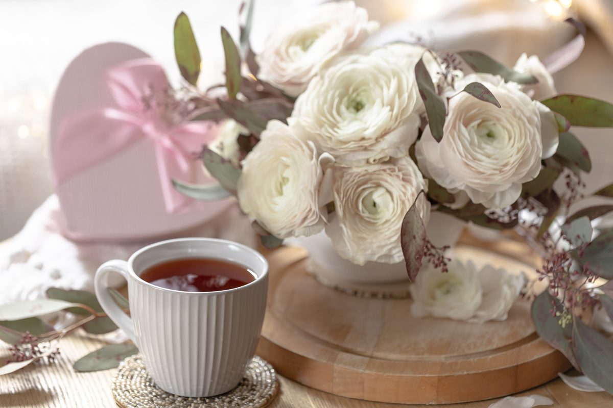 Cup of tea with white ranunculus flowers and a pink heart box on a wooden tray