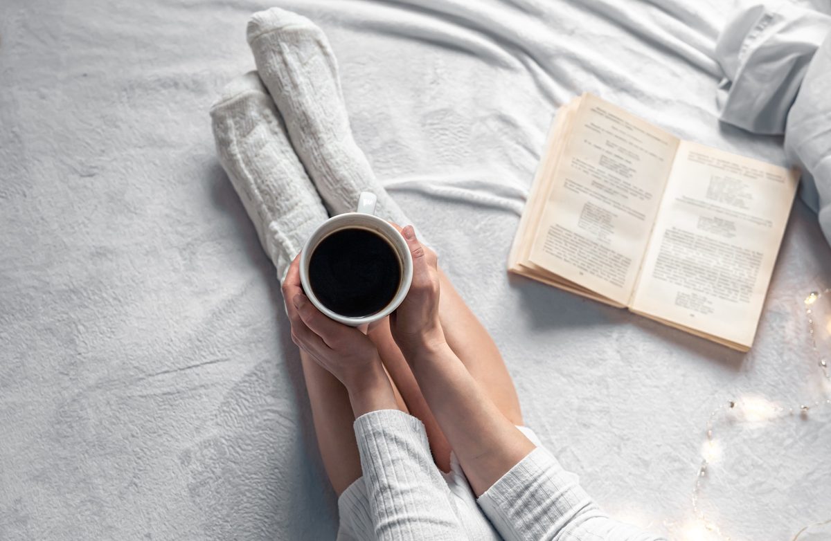 Woman's hands holding a cup of coffee on a cozy bed next to an open book