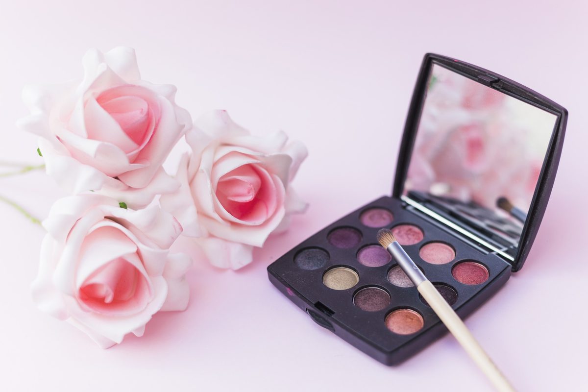 Eyeshadow palette with a mirror, a brush and pink roses on a pink background
