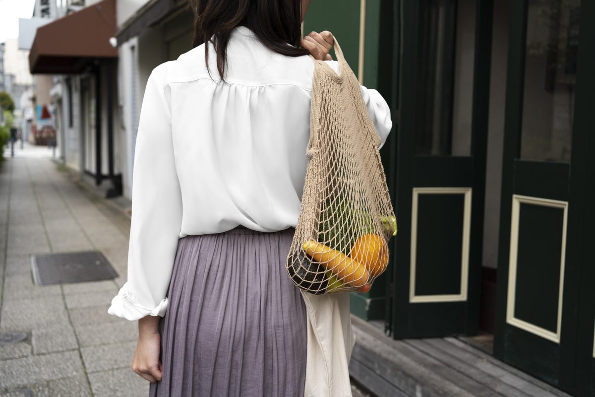 Woman seen from behind walking through a sunny farmers market in a cream sweater and light wash jeans with a woven tote