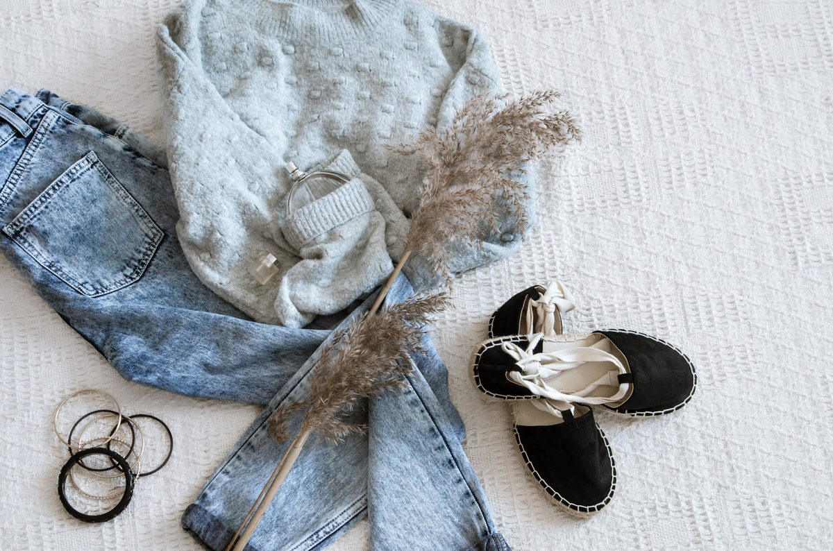 Women's casual weekend outfit flat lay with a cream knit sweater, light wash jeans, white sneakers and gold hoop earrings