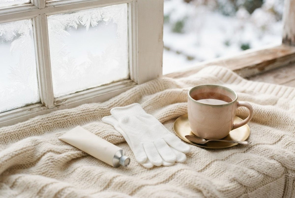 Tube of hand cream, white cotton gloves and a cup of tea on a cream knit blanket by a frosty window